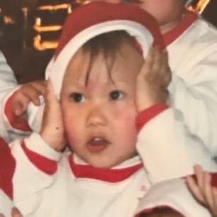 My childhood Christmas portrait, wearing a Santa hat with my hands beside my face.